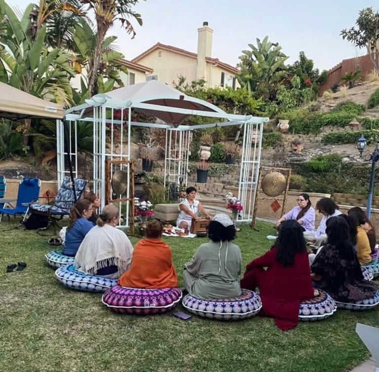 Group sitting in circle on grass facing a gazebo in a backyard setting with a house and palm trees in background